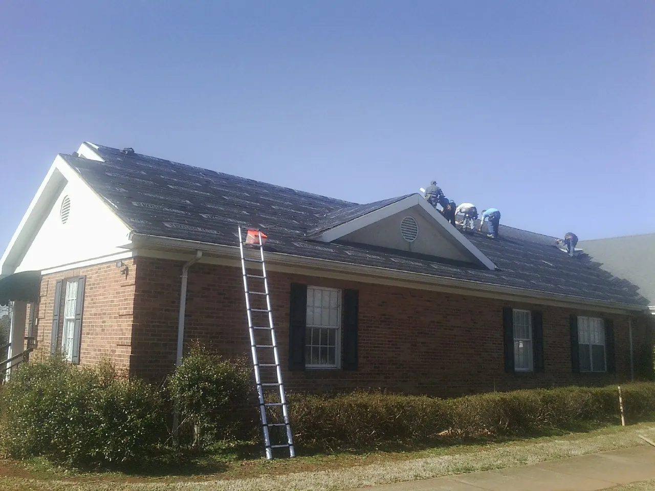 Crew tearing shingles off a brick ranch roof with a ladder set — Metal Roof Repair work by our crew in Hawkeye