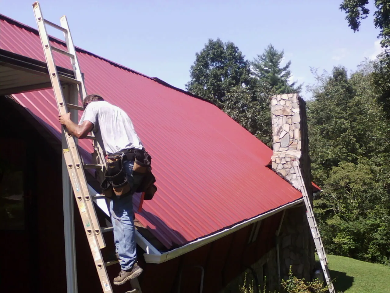 Roofer on a ladder beside a red metal roof and stone chimney — Soffit & Fascia Repair by Roofing Brothers in Hawkeye